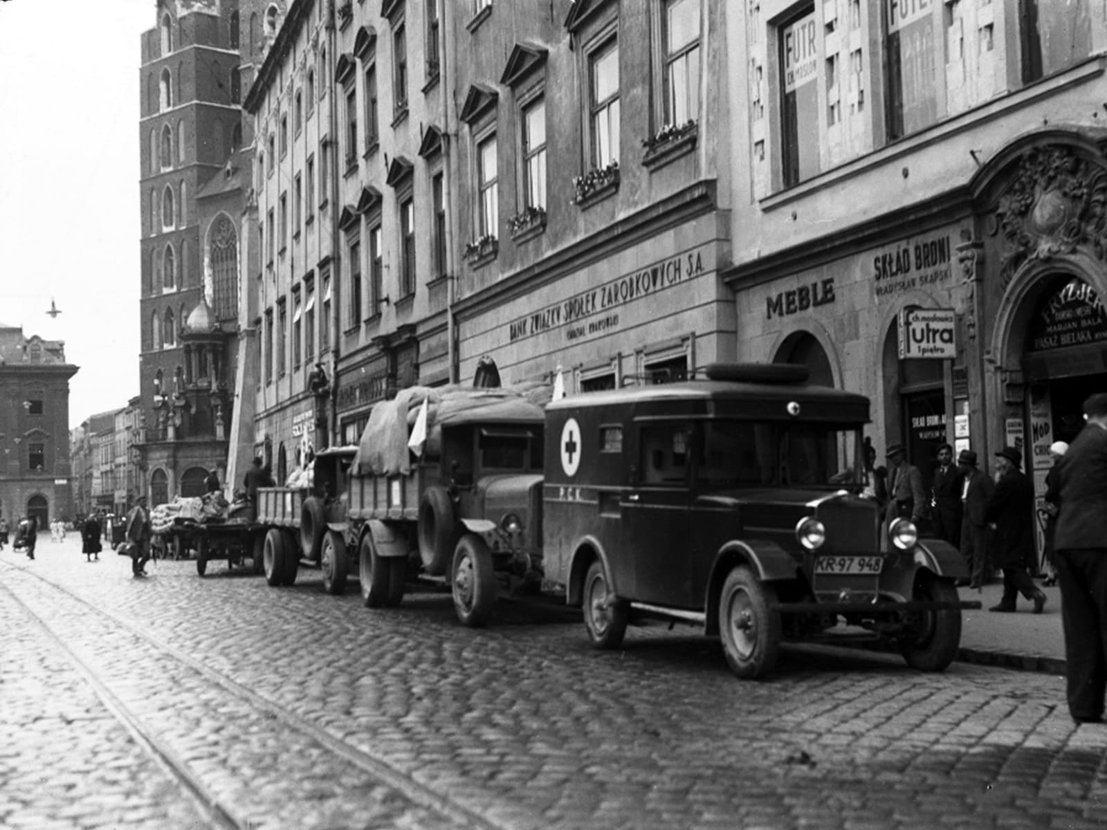 Wyjazd kolumny sanitarnej krakowskiego oddziału PCK do Szczucina na tereny objęte powodzią. Samochody stojące przed Kamienicą Bonerowską w oczekiwaniu na wyjazd. Widoczne reklamy, w głębi kościół Mariacki. Data wydarzenia: 1934-07.